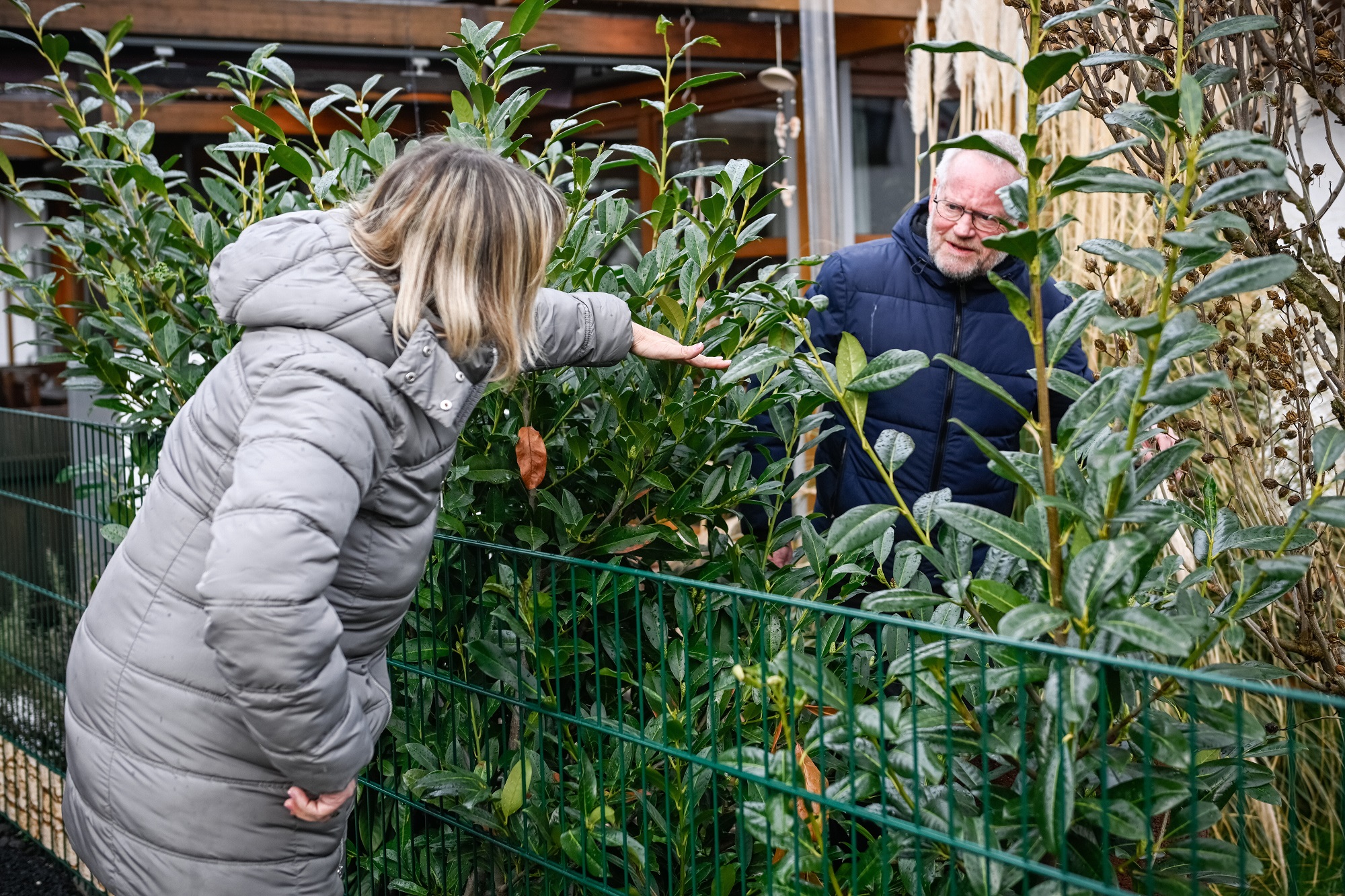 Eine Frau zeigt am Gartenzaun einem Mann auf der anderen Seite die Höhe des Pflanzenbewuchses an.