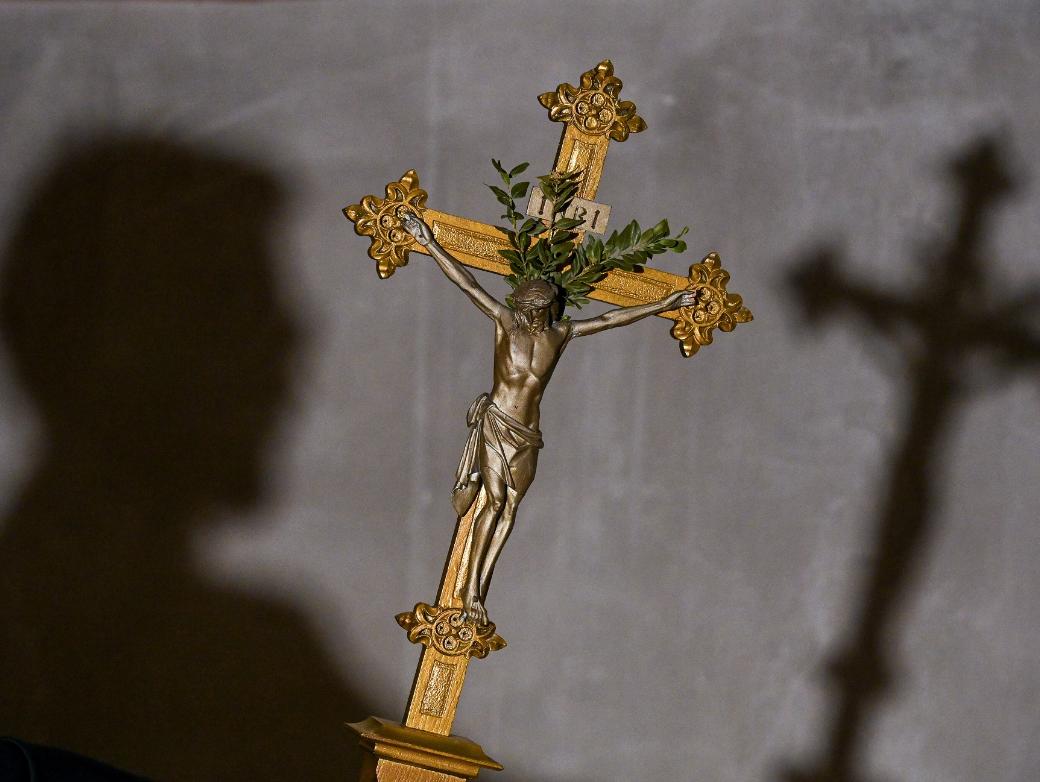 Im Vordergrund des Bildes sieht man ein goldenes Kreuz, im Hintergrund an der Wand den Schatten des Kreuzes und den des Kopfes der Person, die es hält.