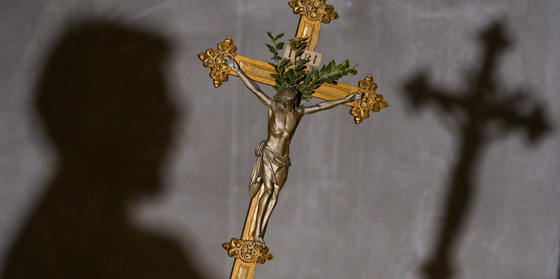 Im Vordergrund des Bildes sieht man ein goldenes Kreuz, im Hintergrund an der Wand den Schatten des Kreuzes und den des Kopfes der Person, die es hält.
