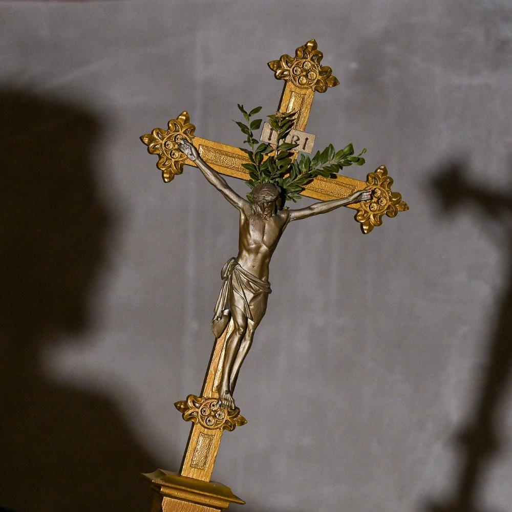 Im Vordergrund des Bildes sieht man ein goldenes Kreuz, im Hintergrund an der Wand den Schatten des Kreuzes und den des Kopfes der Person, die es hält.