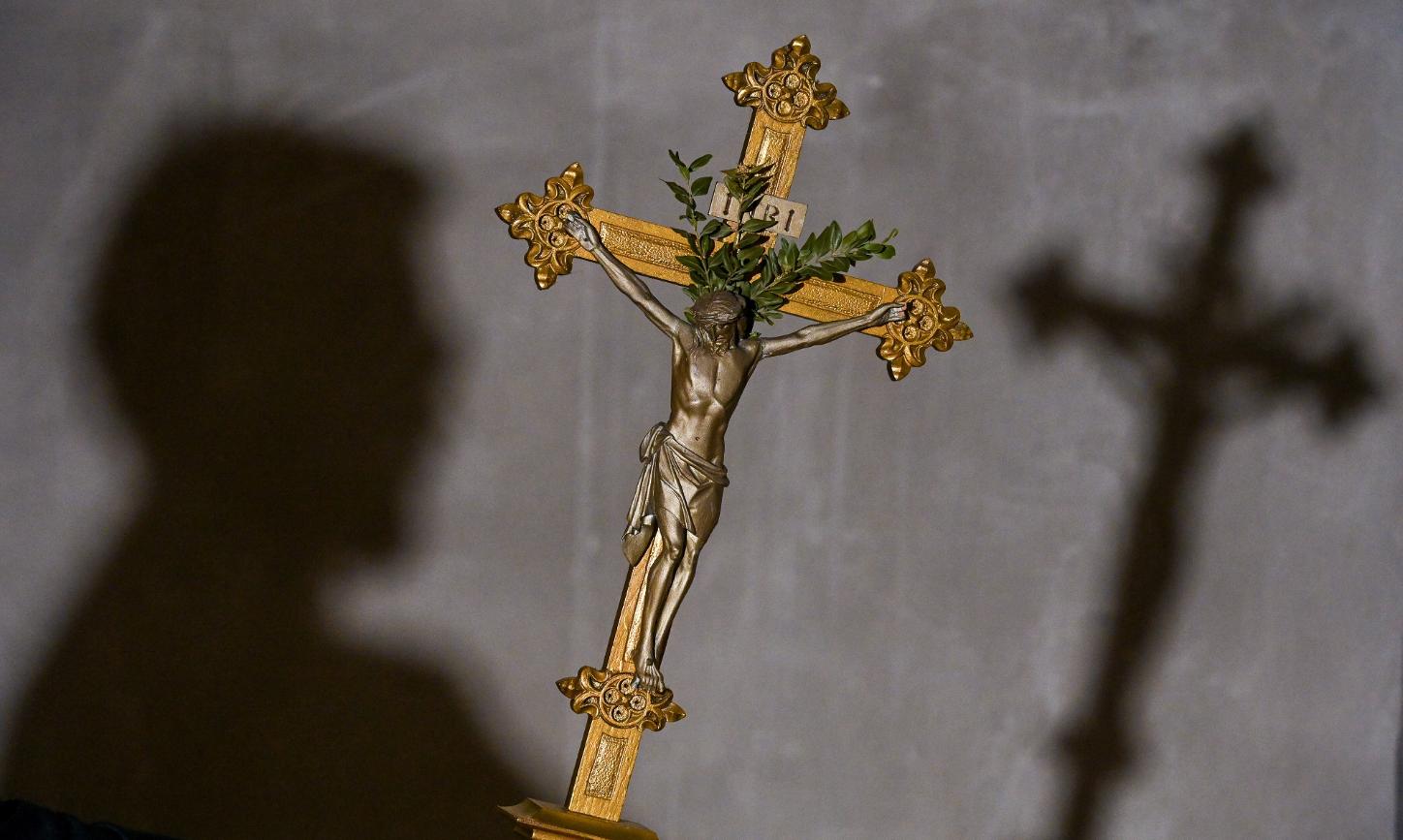 Im Vordergrund des Bildes sieht man ein goldenes Kreuz, im Hintergrund an der Wand den Schatten des Kreuzes und den des Kopfes der Person, die es hält.