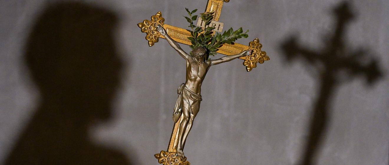 Im Vordergrund des Bildes sieht man ein goldenes Kreuz, im Hintergrund an der Wand den Schatten des Kreuzes und den des Kopfes der Person, die es hält.