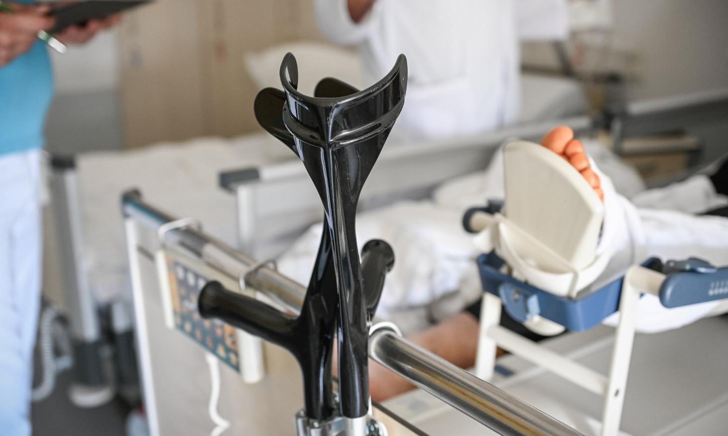An einem Krankenhausbett lehnt ein Paar Krücken. Von der Person im Bett ist ein verletzter Fuß im Bild.
