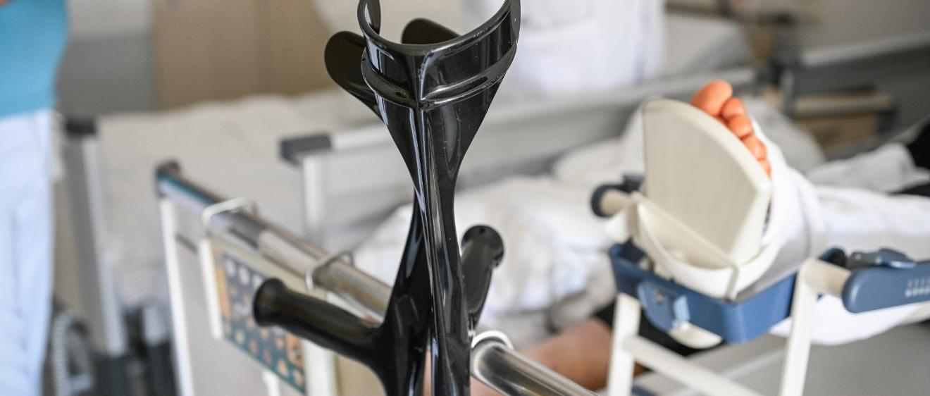 An einem Krankenhausbett lehnt ein Paar Krücken. Von der Person im Bett ist ein verletzter Fuß im Bild.