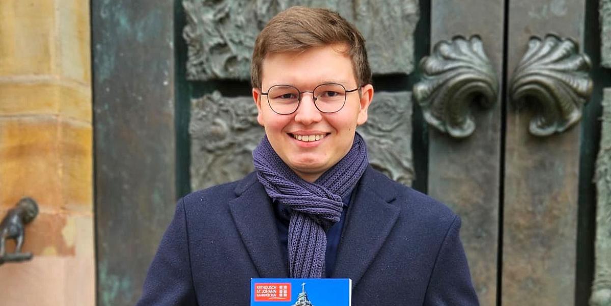 Der 19-jährige David Sawas mit seiner Festschrift über die Saarbrücker Basilika St. Johann vor dem Portal der Kirche, das die künstlerische Handschrift des Bildhauers Ernst Alt trägt. (c) Andreas Kossmann Der 19-jährige David Sawas mit seiner Festschrift über die Saarbrücker Basilika St. Johann vor dem Portal der Kirche, das die künstlerische Handschrift des Bildhauers Ernst Alt trägt.
