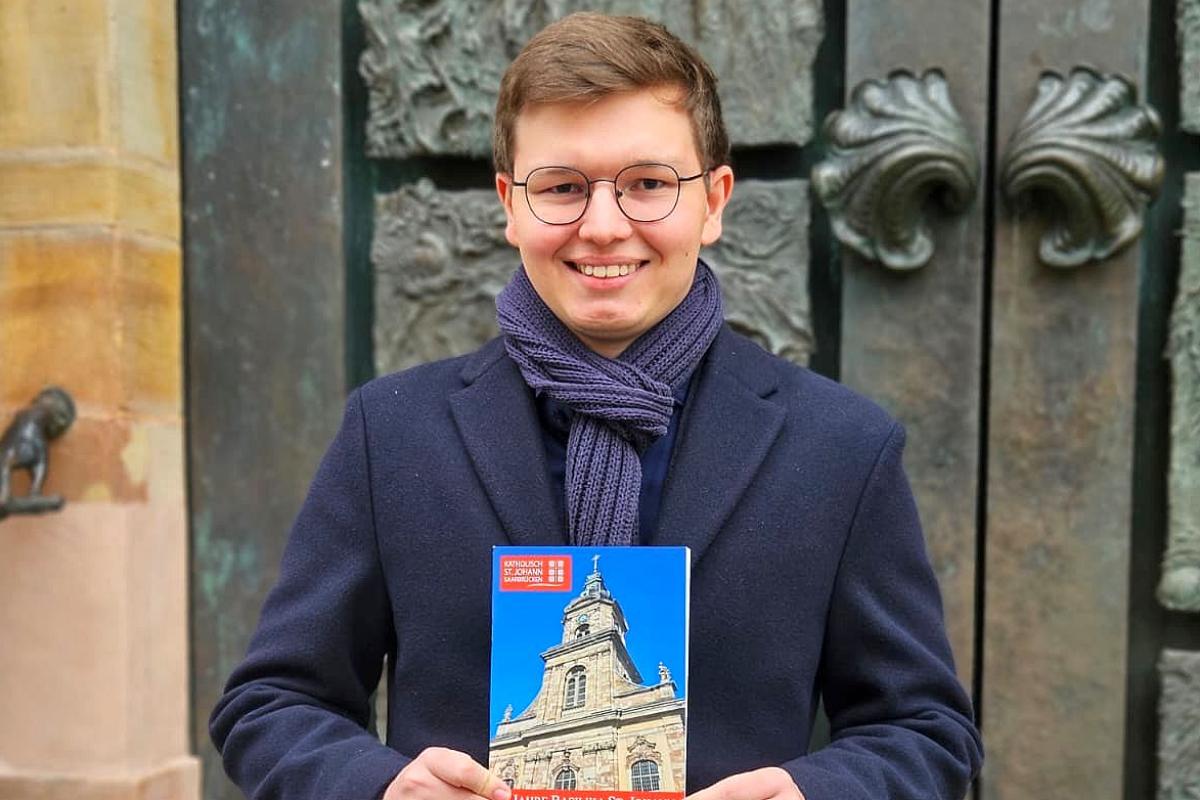 Der 19-jährige  David Sawas mit seiner Festschrift über die Saarbrücker Basilika St. Johann vor dem Portal der Kirche, das die künstlerische Handschrift des Bildhauers Ernst  Alt trägt.