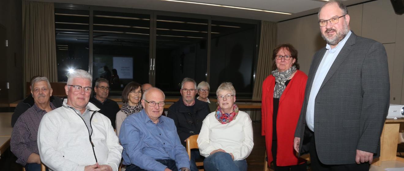 Dekan Jörg Schuh und Pastoralreferentin Susanne Moeckel-Lamberty neben einer Gruppe sitzender Menschen stehend.