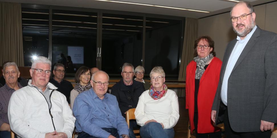 Dekan Jörg Schuh und Pastoralreferentin Susanne Moeckel-Lamberty neben einer Gruppe sitzender Menschen stehend.