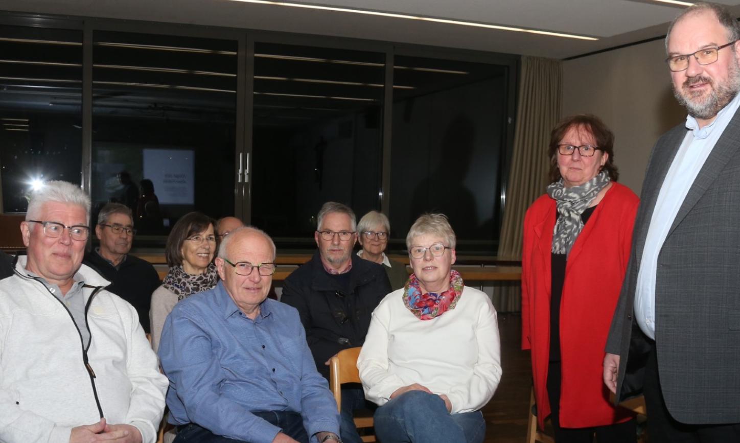 Dekan Jörg Schuh und Pastoralreferentin Susanne Moeckel-Lamberty neben einer Gruppe sitzender Menschen stehend.