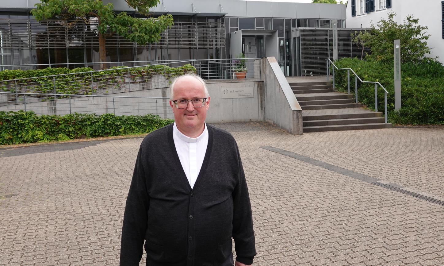 Ein Mann lächelt in die Kamera, im Hintergrund ein großes graues Gebäude