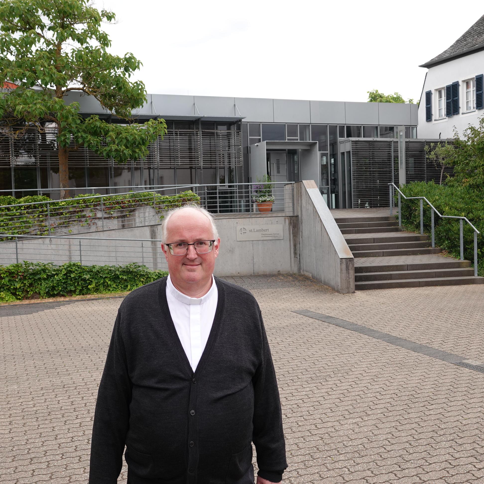Ein Mann lächelt in die Kamera, im Hintergrund ein großes graues Gebäude