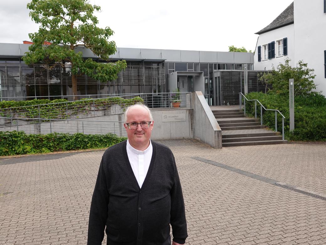 Ein Mann lächelt in die Kamera, im Hintergrund ein großes graues Gebäude