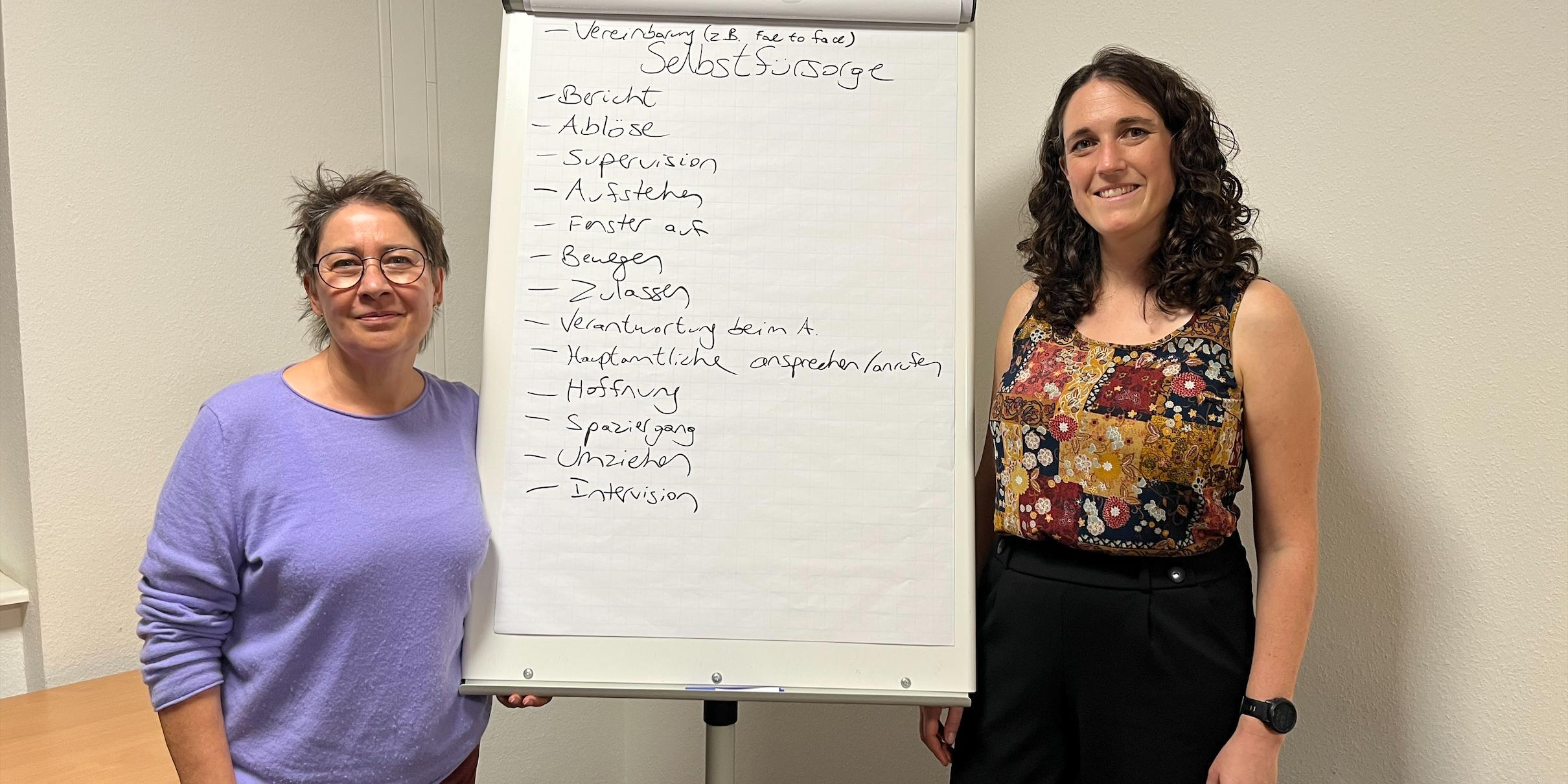 Stephanie Schneider (links), Leiterin der TS Trier, und Psycho login Dr. Jessica Levy geben den Ehrenamtlichen Tipps für den Umgang mit Suizidgedanken am Telefon. (c) Sarah Andre-Schött Zwei Frauen blicken in die Kamera, zwischen ihnen eine Pinnwand mit Notizen