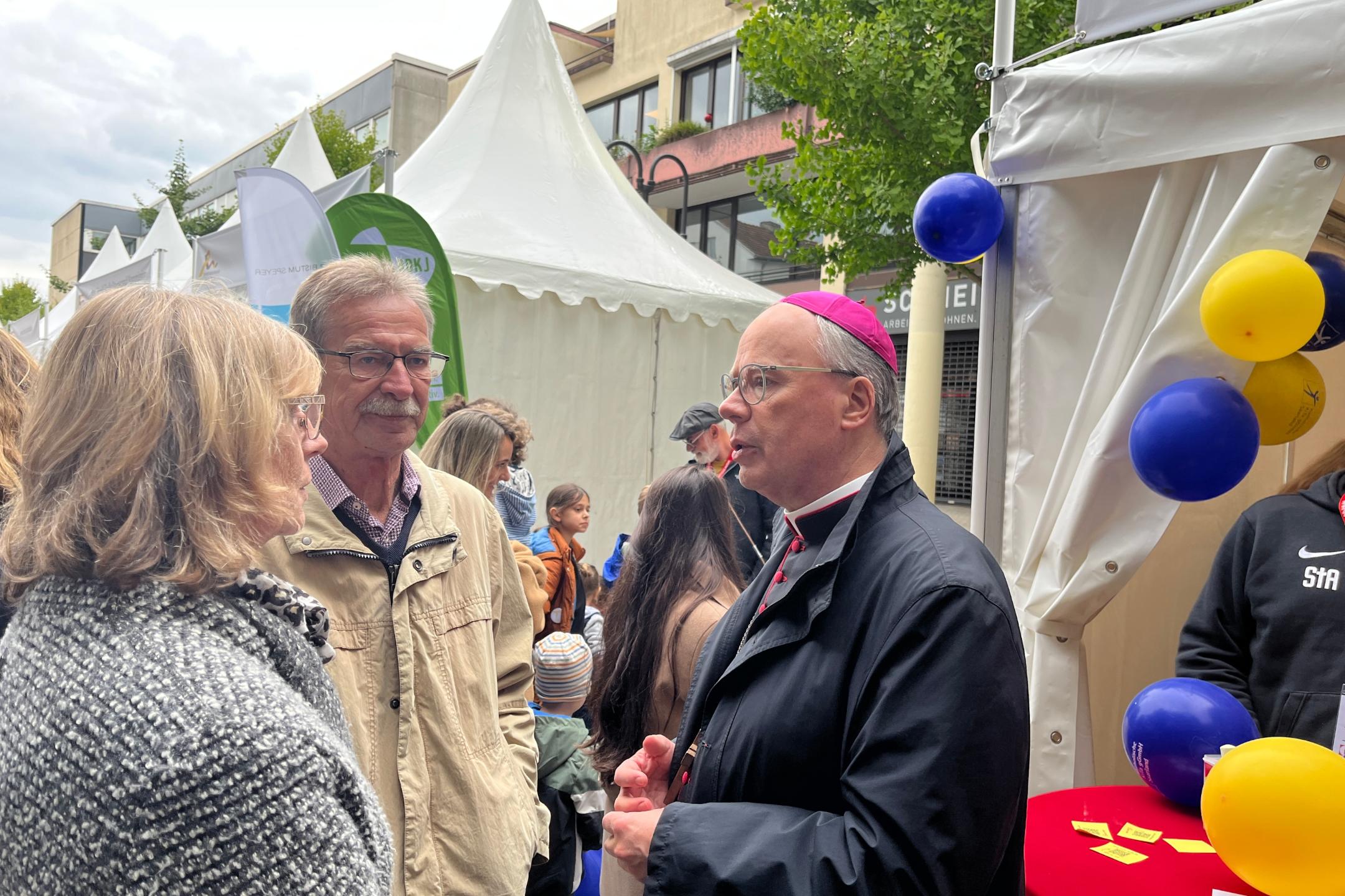 Ein Bischof spricht mit einem Mann und einer Frau vor einem Zelt mit Luftballons