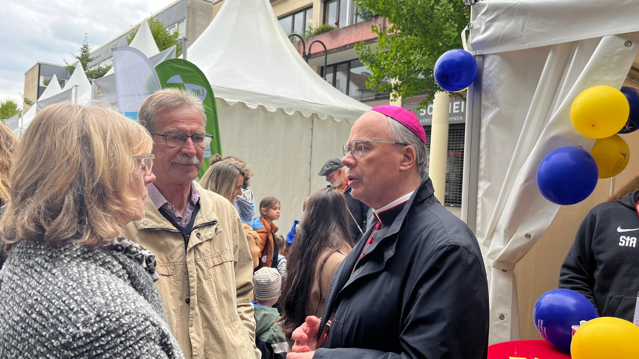 Ein Bischof spricht mit einem Mann und einer Frau vor einem Zelt mit Luftballons
