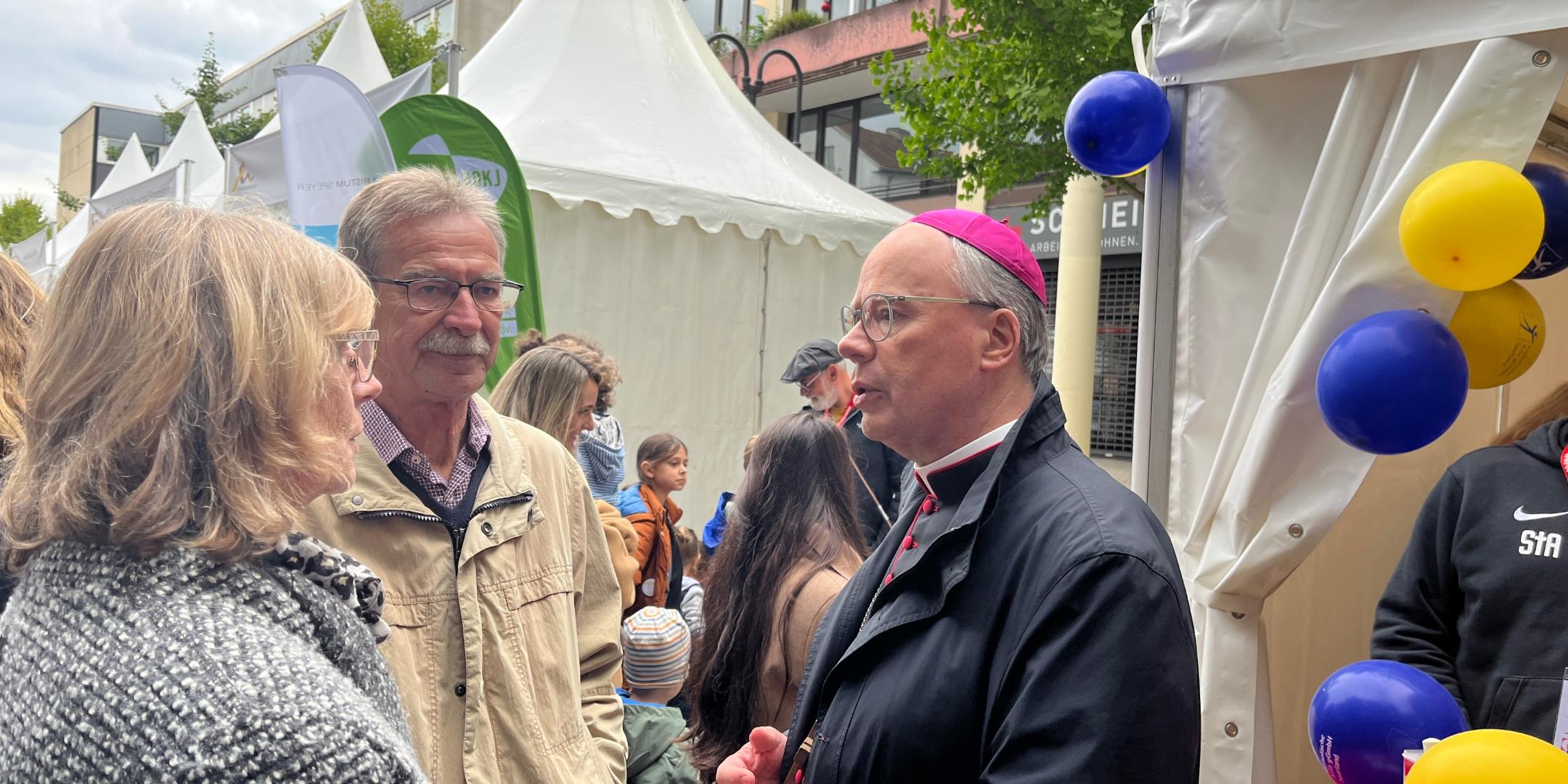 Ein Bischof spricht mit einem Mann und einer Frau vor einem Zelt mit Luftballons (c) Ute Kirch/Bistum Ein Bischof spricht mit einem Mann und einer Frau vor einem Zelt mit Luftballons