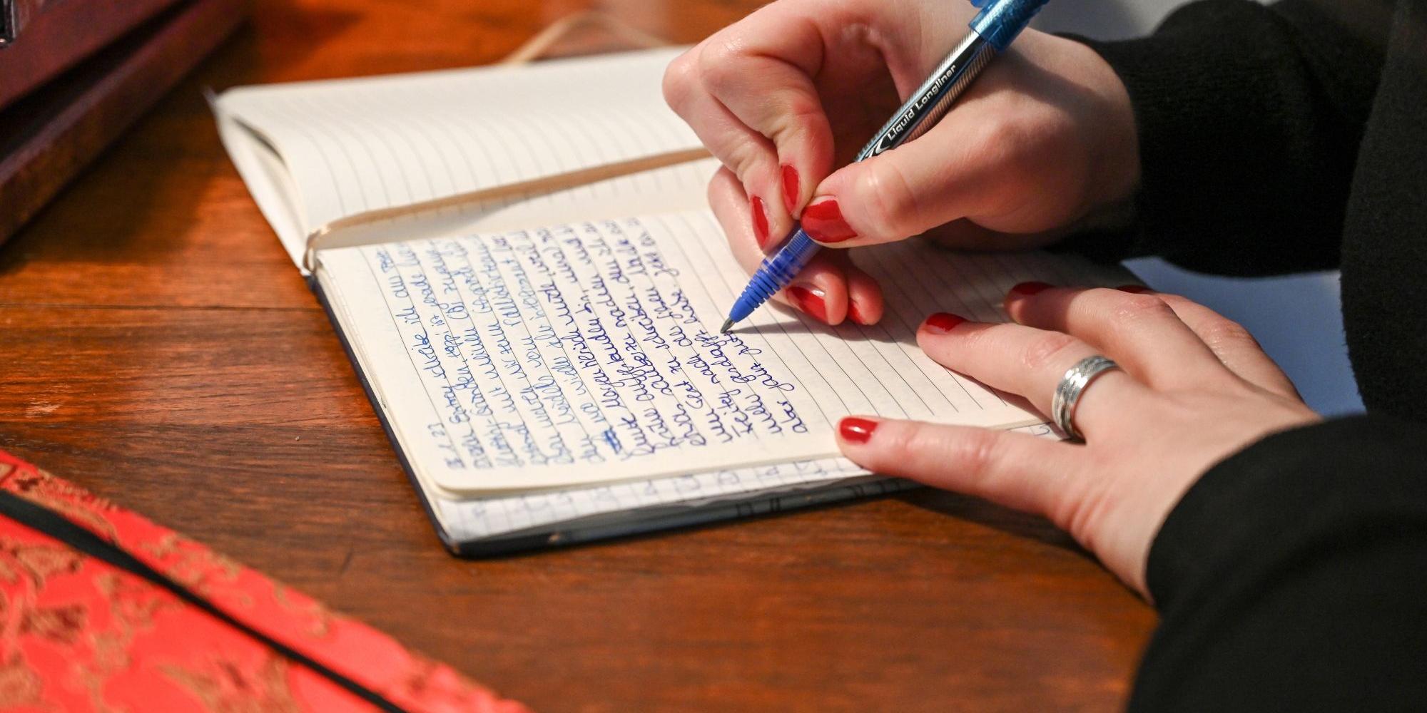 Eine Frau schreibt in ihr Tagebuch, vor ihr liegen weitere Kladden.