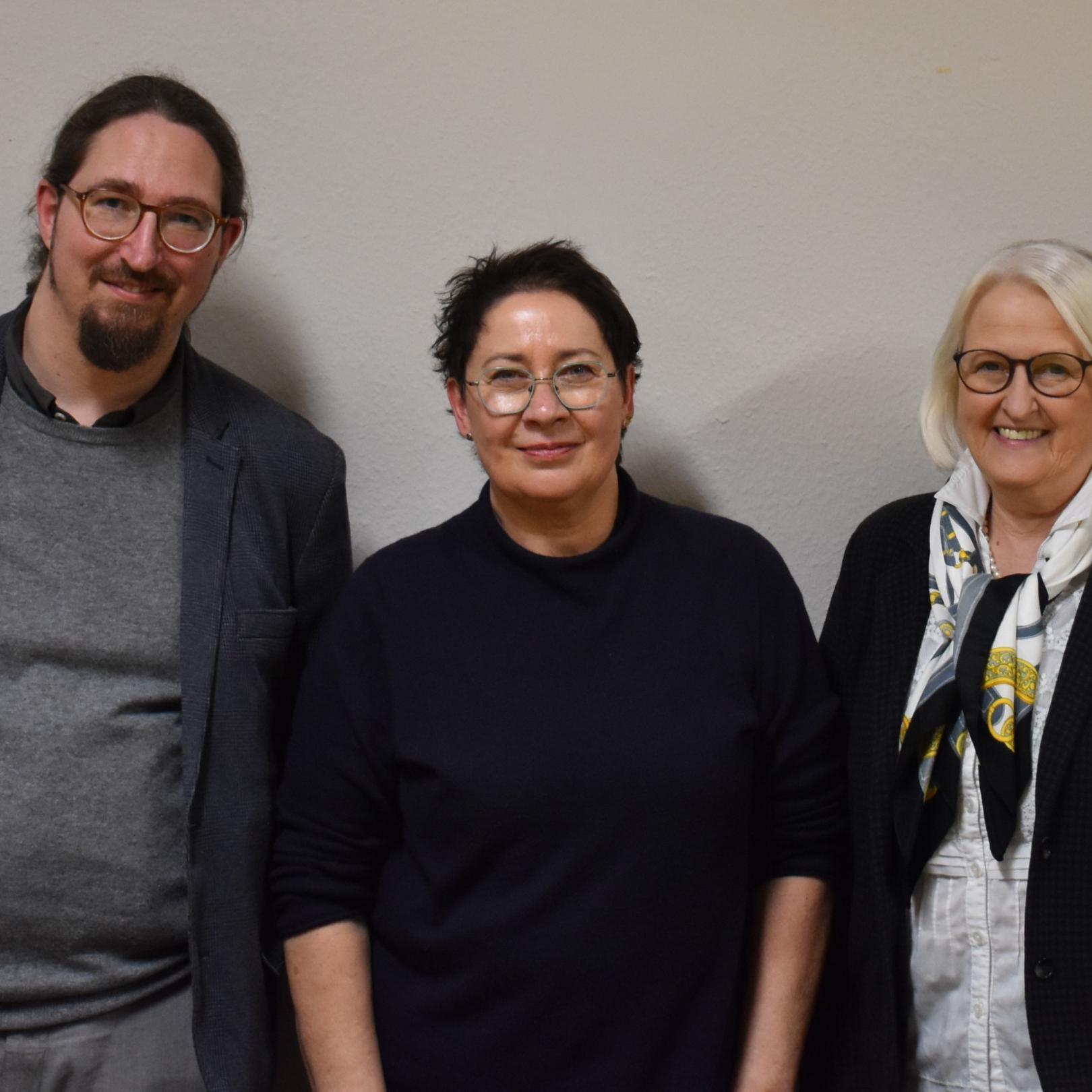 Ein Mann und zwei Frauen vor einer weißen Wand, alle dunkel gekleidet