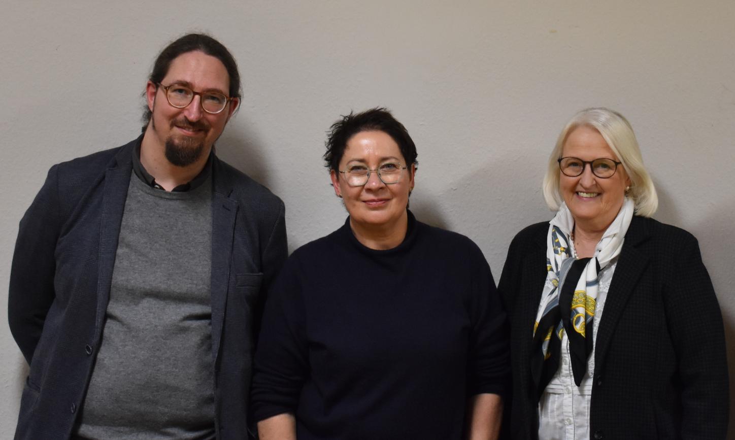 Ein Mann und zwei Frauen vor einer weißen Wand, alle dunkel gekleidet