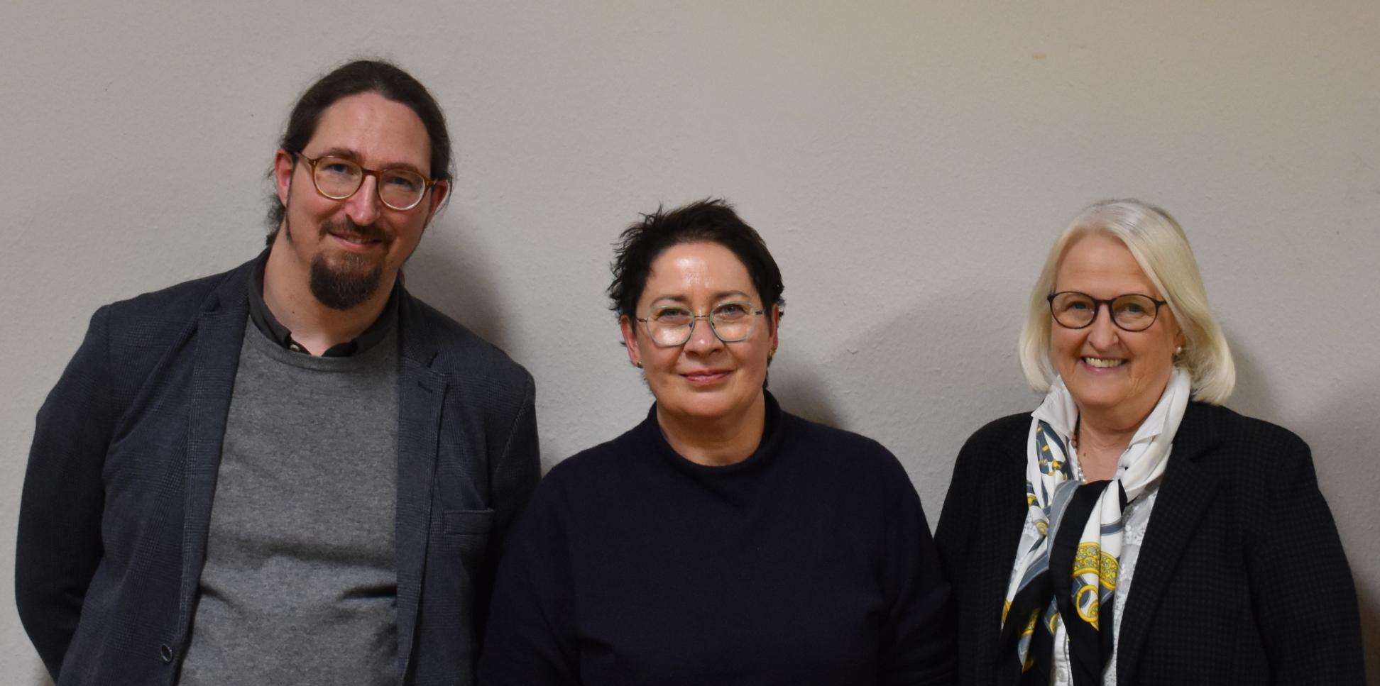 Ein Mann und zwei Frauen vor einer weißen Wand, alle dunkel gekleidet