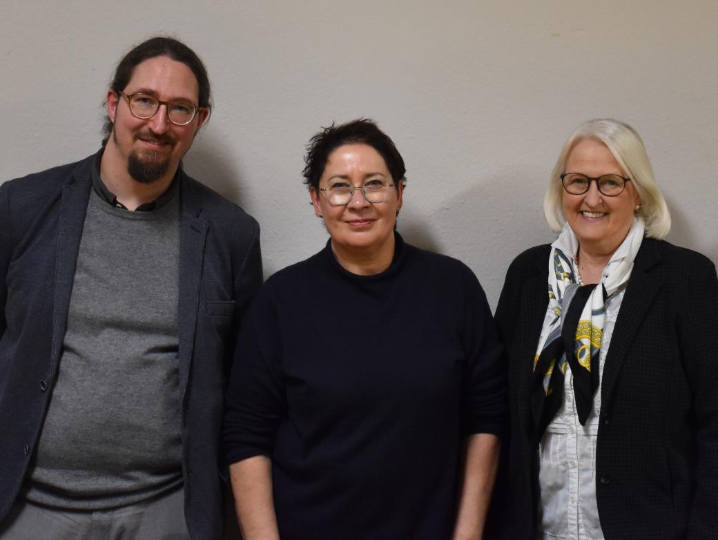 Ein Mann und zwei Frauen vor einer weißen Wand, alle dunkel gekleidet