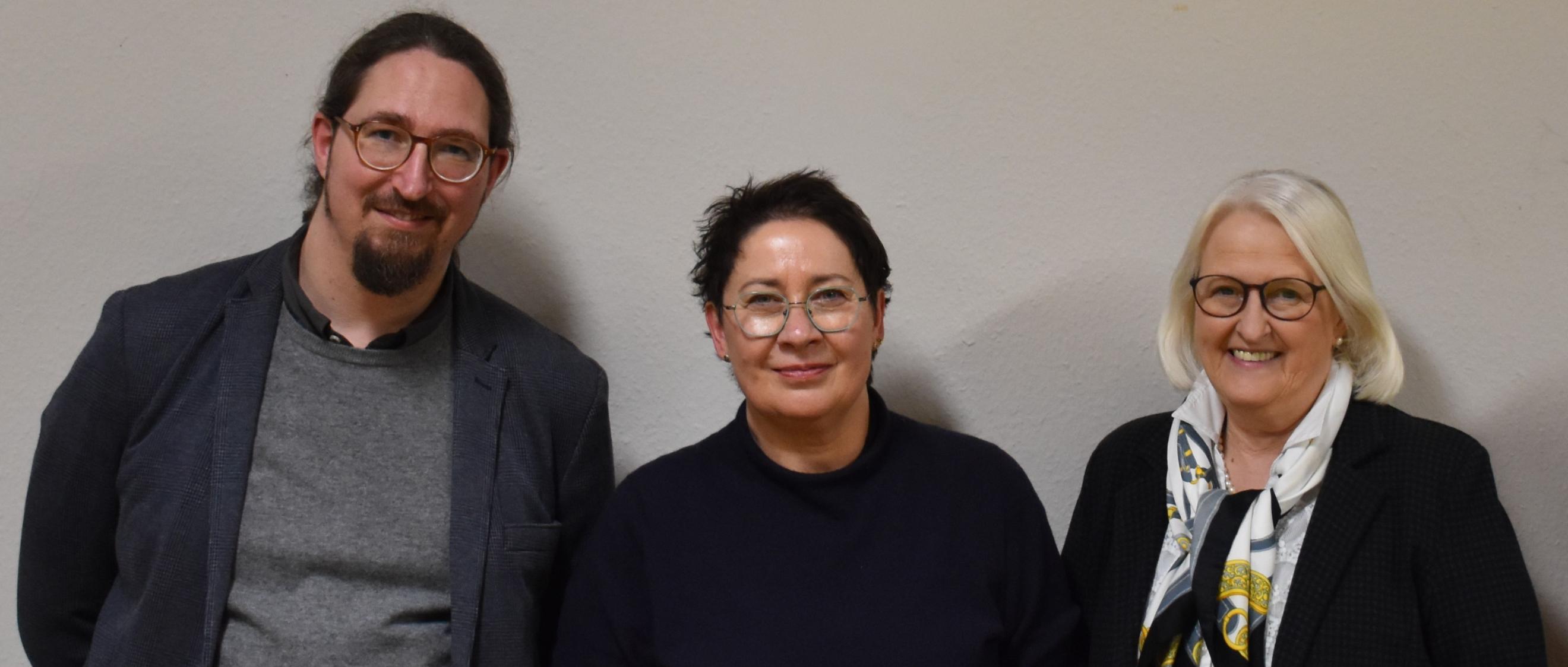 Ein Mann und zwei Frauen vor einer weißen Wand, alle dunkel gekleidet