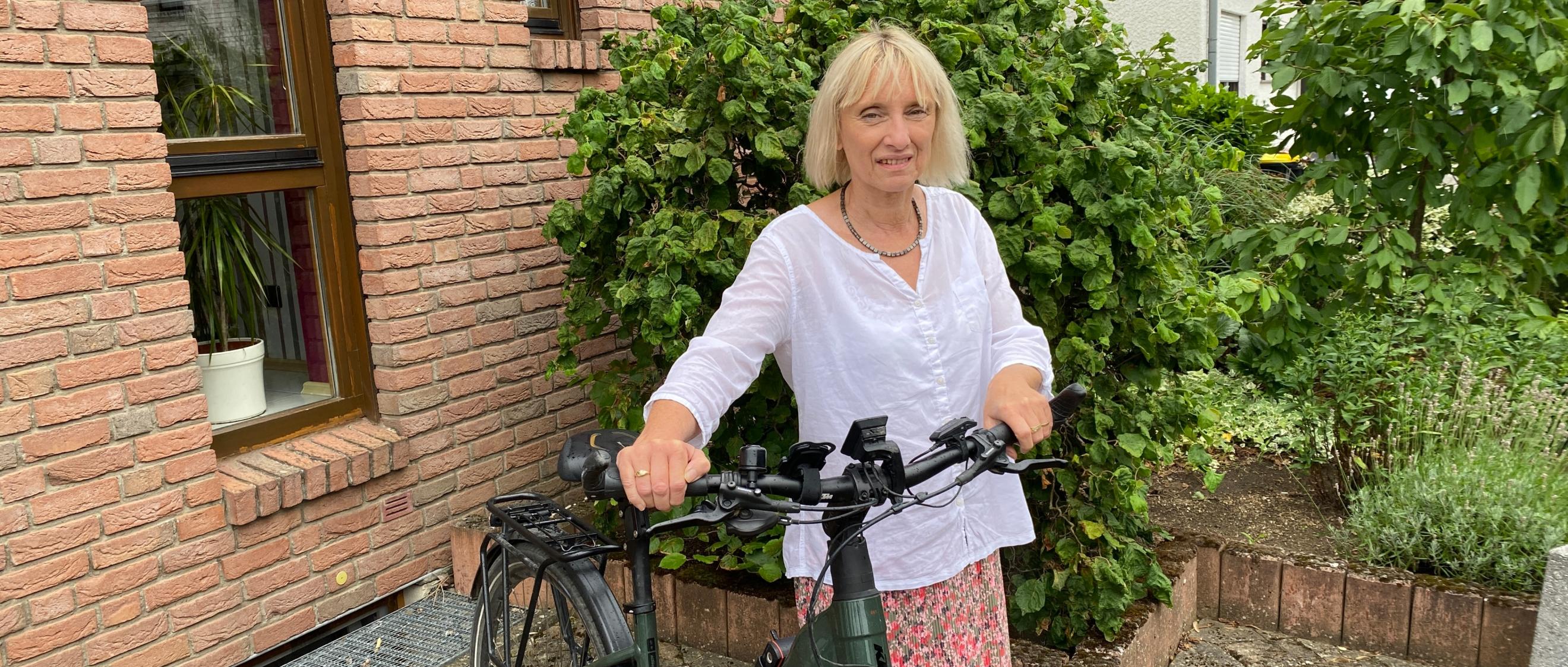 Eine Frau mit Fahrrad vor einem Backsteinhaus blickt lächelnd in die Kamera.