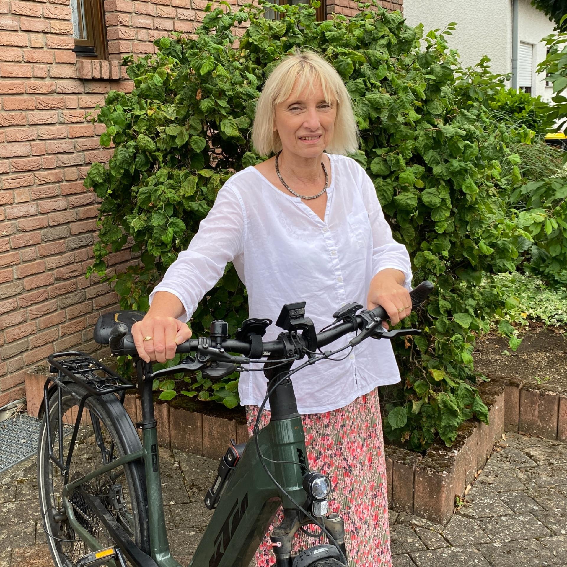 Eine Frau mit Fahrrad vor einem Backsteinhaus blickt lächelnd in die Kamera.