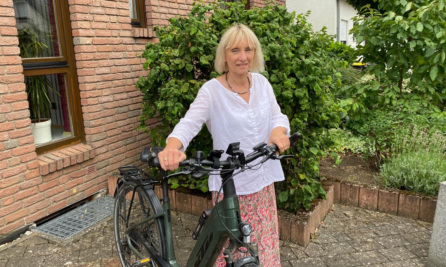 Eine Frau mit Fahrrad vor einem Backsteinhaus blickt lächelnd in die Kamera.