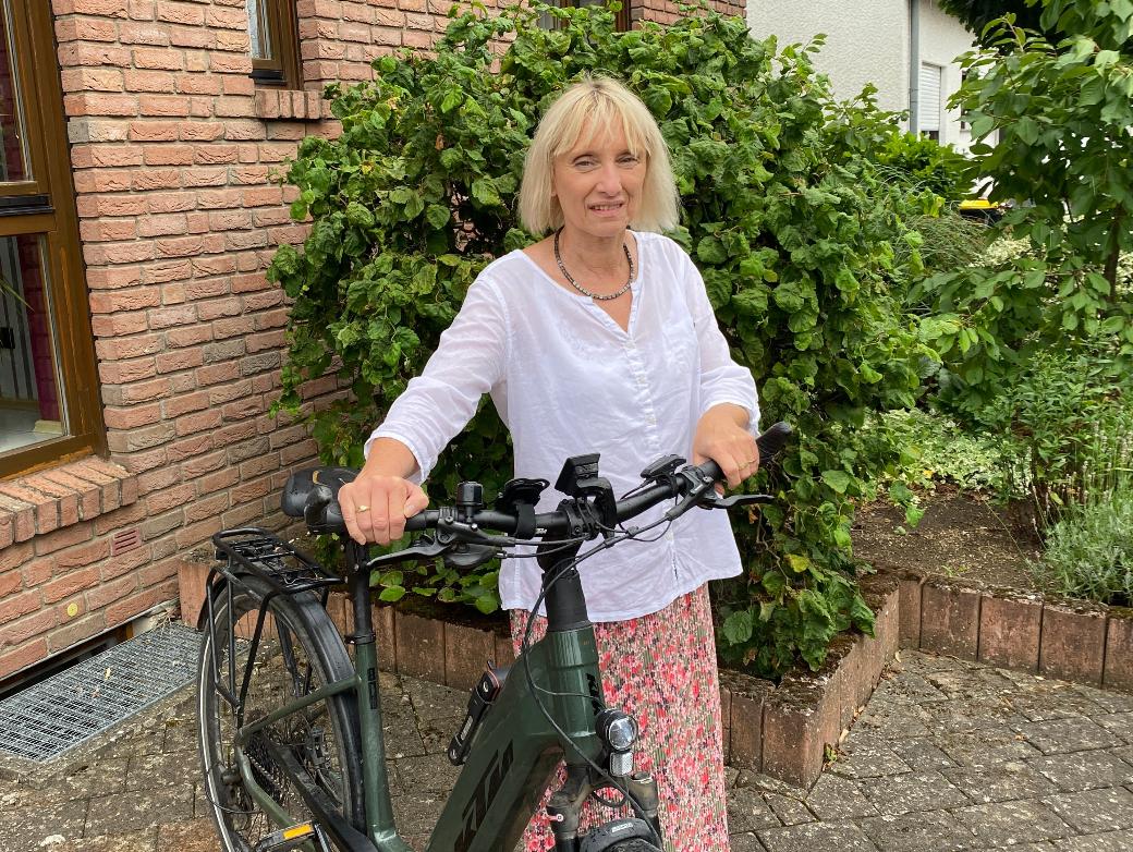 Eine Frau mit Fahrrad vor einem Backsteinhaus blickt lächelnd in die Kamera.