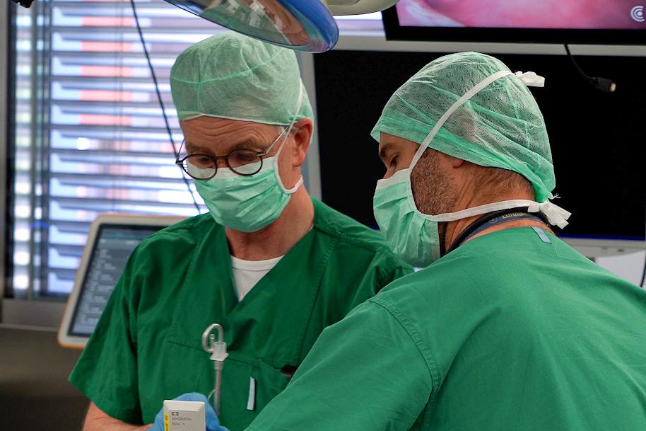 Zwei Männer in Grünen Hemden mit Mundschutz arbeiten an einem Patienten