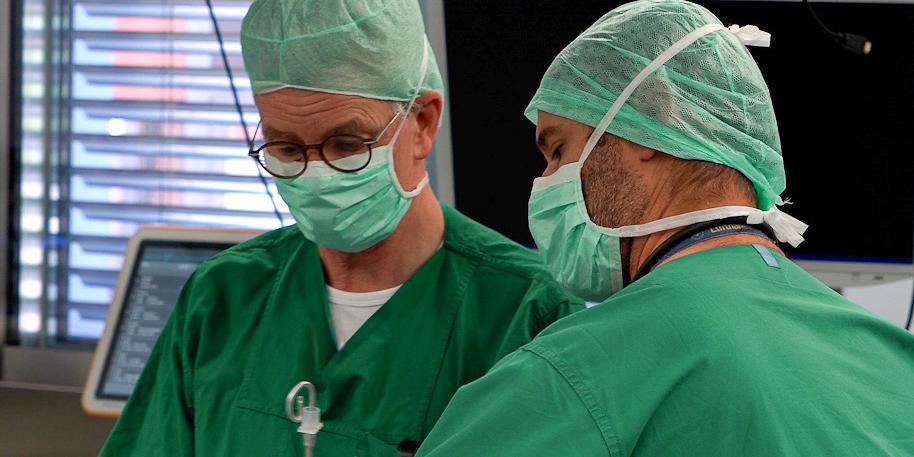 Zwei Männer in Grünen Hemden mit Mundschutz arbeiten an einem Patienten