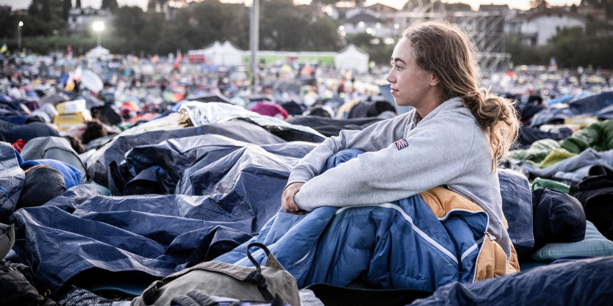 Am Morgen nach der Gebetsnacht beim Weltjugendtreffen 2025 in Rom: Eine junge Frau sitzt in ihrem Schlafsack und blickt über das Gelände.