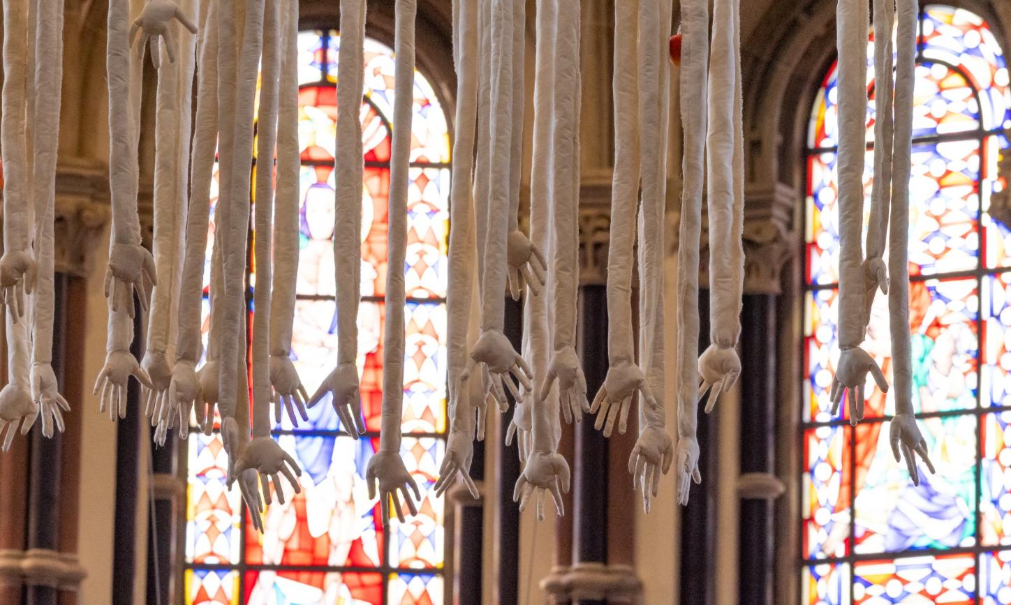 Das Kunstwerk „Mensch“ von Barbara Gröbl zeigt Arme und Hände, die vom Kirchengewölbe herabhängen, geschaffen aus Leinen, mit Äpfeln.
