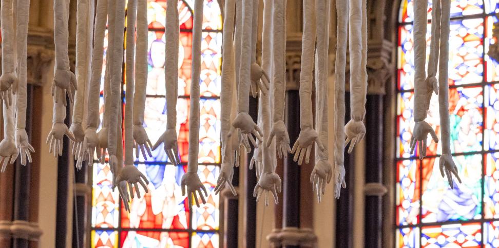 Das Kunstwerk „Mensch“ von Barbara Gröbl zeigt Arme und Hände, die vom Kirchengewölbe herabhängen, geschaffen aus Leinen, mit Äpfeln.