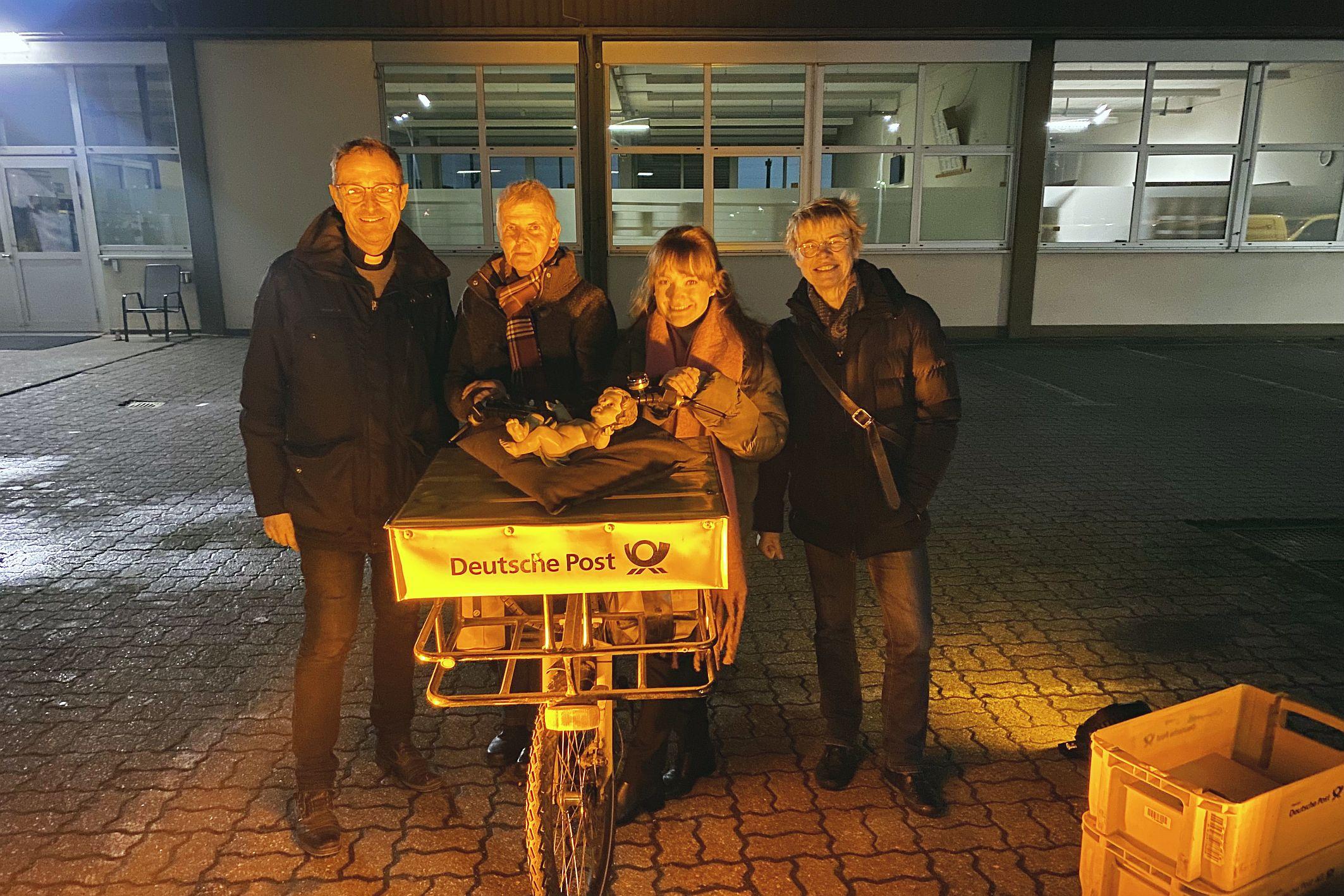 Pfarrer Jörg Meyrer, Ulrike Phiesel, Nadine Kreuser und Bärbel Knieps vom Vor­bereitungsteam mit der Figur des Jesuskinds, das von einem Postboten per Fahrrad geliefert wurde.